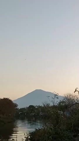The video shows a serene landscape with a mountain in the distance. A narrow river flows through the scene, bordered by lush greenery and trees. The sky is a pale, clear blue, indicating it could be early morning or late afternoon. The overall mood is peaceful and tranquil, capturing the beauty of nature.