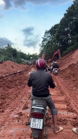 The video shows a muddy, red dirt road, likely after rainfall or a landslide. Several motorcycles are visible, with one in the foreground from a rider's perspective. In the distance, a large crowd of people are gathered, and heavy construction equipment, specifically excavators, are present, suggesting ongoing work or a response to an event. The environment is rural, with trees lining the sides of the road and a cloudy sky. The road surface is heavily rutted and appears difficult to traverse.