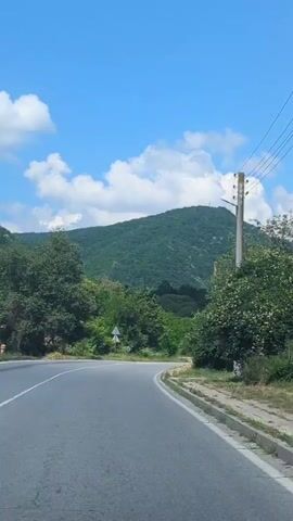 The video shows a road curving through a lush green landscape under a bright blue sky with fluffy white clouds. A white truck is driving away from the camera on the road. In the background, a large, tree-covered mountain dominates the horizon, with a small structure visible at its peak. Trees and bushes line the sides of the road, and a utility pole with wires stands to the right. A small house with a tiled roof and pink flowers is also visible to the right of the road. The scene appears to be set in a rural or mountainous area, possibly Pchelinovo, Bulgaria, on a sunny day. There are no people visible in the video, and the overall mood is peaceful and scenic. The video does not contain any visible text.