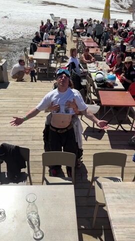 The video features a lively outdoor scene on a sunny day at a ski resort in Saint-Chaffrey, France. The primary subject is a man standing on a wooden deck, wearing a white cropped t-shirt with "Aries" visible, black pants, a brown belt, ski goggles on his forehead, and a bandana. Tattoos are visible on his abdomen, including "Love always-", and abstract designs on his arms. Another person stands behind him, holding up his t-shirt, exposing his midriff. The man smiles broadly with his arms outstretched. He has small hoop earrings, and the person behind him appears to be laughing or shouting.

The wooden deck is furnished with numerous tables and chairs, where many people are seen dining, drinking, and socializing. In the background, snow-covered mountains, ski slopes, and ski lift infrastructure are clearly visible, indicating a bustling ski resort. Ski equipment is stacked near a red flag. The man eventually sits down at one of the tables. The atmosphere is cheerful and relaxed under bright sunshine.