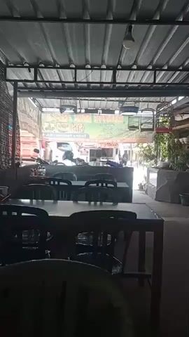The video shows an interior view of a restaurant. In the foreground, there are several tables and chairs. The tables are dark-colored and appear to be made of wood. The chairs are black and made of plastic. The space is open-air, with a corrugated metal roof supported by metal beams. In the background, there is a street scene visible through an open entrance. Vehicles, including a truck and motorcycles, are present on the street. There are also signs and some potted plants visible. The lighting is dim, with a single bulb visible overhead. The overall impression is of a casual, possibly outdoor dining establishment.