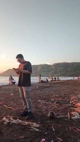 The video shows a beach at sunset. Several people are standing and walking around on the sand. Some are looking at their phones or taking pictures. There is a lot of driftwood scattered on the beach. The sun is setting over the ocean, creating a bright glare. The sky is clear.