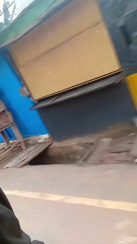 The video shows a journey from the outside of a building into the inside of a yellow motor vehicle with three wheels, commonly known as a "keke."
The video begins by panning across an urban environment with a building covered with blue tarpaulin and wooden structures. A man can be seen sitting near a grey door. There are posters attached to a brick wall and some grass growing in the area. 
The camera moves quickly to the yellow motor vehicle, and the perspective shifts to an interior view looking out through the windshield. The driver is visible, steering the vehicle on a paved road.  Several other vehicles are seen on the road ahead.