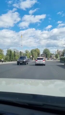 A video taken from inside a car shows a view of a road with traffic. A silver car is in front, followed by a bus and other vehicles. A black SUV is visible on the left side of the road, and several other cars are parked. Trees and buildings line the street, and a Ukrainian flag flies on a pole in the distance. The sky is blue with white clouds.