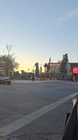 The video shows a street view from a pedestrian perspective, focusing on an intersection during daylight. The sky is clear with a hint of sunset hues. A woman with long, dark curly hair is standing on the sidewalk near the curb, partly obscuring the view. Cars and people move across the intersection as traffic signals change. In the background, buildings are visible, with one displaying a large digital billboard cycling through McDonald's advertisements. A Chilean flag is visible on a building farther back. The overall atmosphere is that of an urban street scene at dusk.