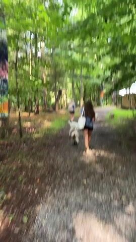 The video shows a dirt path through a wooded area. Two young women are walking away from the camera on the path, each with a small goat walking alongside them. The woman in the foreground has long, dark, wavy hair and is wearing a dark shirt and shorts, carrying a white bag. The woman in the background has lighter hair and is wearing a light-colored shirt and dark shorts. The goats are white and light brown. The path is lined with trees and foliage. To the right of the path, there is a small structure and some barrels. There are also colorful painted poles or totems visible in the background. The overall scene is one of a casual walk or hike in a natural setting.
