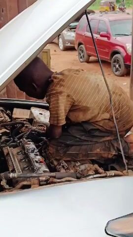 The image depicts an outdoor mechanic's workshop in Karshi, Nigeria, under clear daylight conditions. In the foreground, a young male, appearing to be an apprentice or mechanic, is deeply engrossed in working on the engine of a light-colored (possibly silver or white) car.

The car's hood is fully open, revealing an oily and dirty engine compartment. The young man, dressed in a soiled light-brown and white striped short-sleeved shirt and dark, equally dirty trousers, is hunched over, leaning into the engine bay with his hands inside, manipulating various components. His posture suggests intense focus and concentration on the task at hand, which appears to be engine repair or maintenance.

The background shows an unpaved, reddish-brown dirt ground characteristic of an informal workshop setting. Other vehicles are visible; immediately behind the main subject's car is a silver-grey car with its hood also open. Further to the right, a red SUV is parked. Distant structures and some sparse vegetation are visible, suggesting an open-air environment. No discernible text is visible on any objects within the frame. The overall scene conveys a busy, hands-on repair operation.