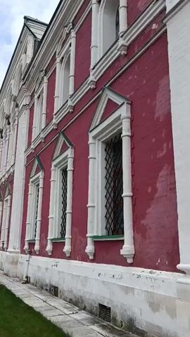 The video pans across the exterior of a large, old building with a pinkish-red brick facade and white decorative trim. The building has tall, arched windows with black iron grilles. A paved walkway runs alongside the building, bordering a garden bed filled with rose bushes and other greenery. The sky is partly cloudy. The camera moves from left to right, showing more of the building and the garden as it progresses. The overall impression is one of an old, well-maintained historical site.