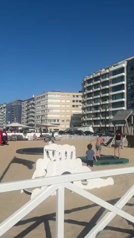 The video shows a sunny day at a sandy playground near a beach with buildings in the background. Three children are visible: two boys and a girl. One boy wears a blue t-shirt, another is shirtless with patterned swim trunks, and the girl wears a black top and shorts. They are near a sunken trampoline and a small wooden playhouse with a slide and swingset. A white fence is in the foreground, partially obscuring the view. Cars are parked along the street behind the playground. A bird flies in the sky. Other people can be seen in the background.