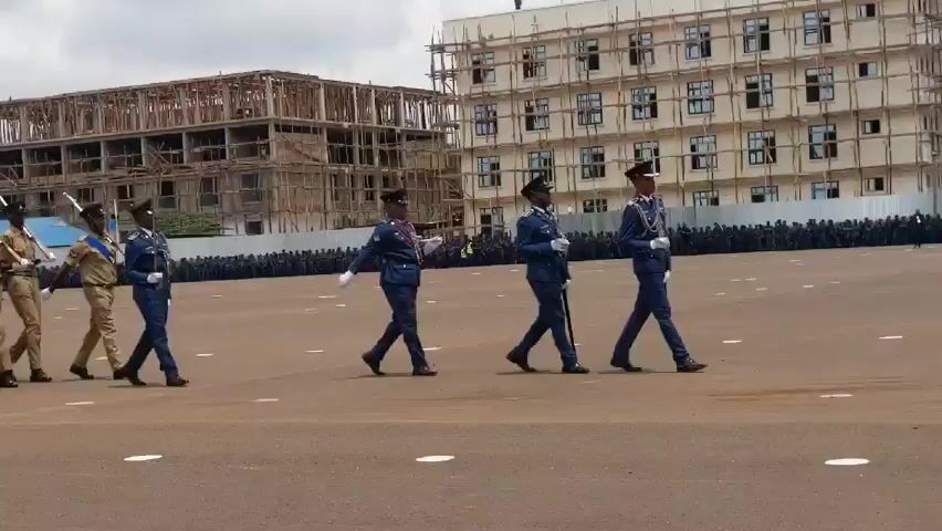 In a wide shot, a column of men in tan uniforms march in formation on a large, brown surface, likely a parade ground. They are holding rifles with bayonets affixed, and their movements are synchronized. Alongside this group, four men in blue uniforms march. A large group of people is visible in the background. In the background, there are two buildings; one appears to be under construction, while the other looks complete. The sky is overcast.
