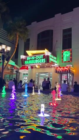 At night, an In-N-Out Burger restaurant is seen behind a fountain of brightly colored water jets. The building is white with a red and green In-N-Out sign. A neon green palm tree sign is adjacent to the restaurant. People are standing and sitting around the fountain and by the restaurant. The lighting is bright and colorful. Other storefronts can be seen nearby.