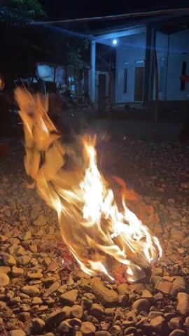 The video presents an outdoor night scene in Meurinya Tanjong, Indonesia, centered around a vibrant bonfire. Bright orange and yellow flames dance vigorously, casting a warm glow over a ground covered in small, light-colored pebbles or gravel.

In the mid-ground, a modern-style house is partially visible, illuminated by a distinct blue light emanating from its porch or entrance area. The house features clean architectural lines.

Several individuals are present in the background. On the right side of the frame, a person, likely a child or young adult wearing a light-colored shirt, stands partially obscured, observing the fire. Faint silhouettes of other people can be discerned further in the distance on the left side of the frame.

The overall setting suggests a casual, informal gathering, possibly for warmth, light, or an outdoor activity. The time of day is clearly night, with the bonfire and the house lights providing the main sources of illumination. There is no visible text within the frame. The video primarily focuses on the dynamic and captivating movement of the large bonfire.