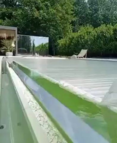 The video shows a close-up of a green swimming pool covered by a white, transparent, corrugated plastic sheeting. The sheeting is partially rolled back, revealing the water underneath. The pool is surrounded by lush green trees and bushes, and in the background, a white lounge chair is visible next to the pool. The scene appears to be a sunny day.