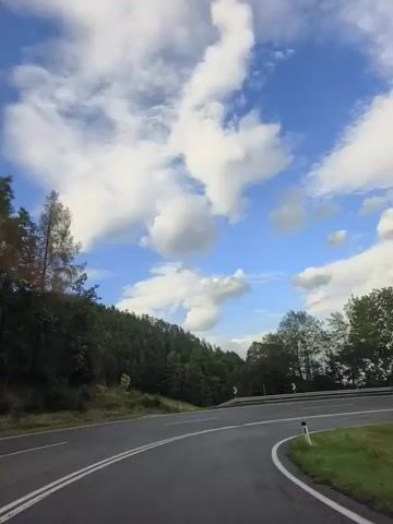 The video shows a scenic view from a moving vehicle on a winding road. The sky is blue with scattered white clouds. Lush green trees and a forested hill are visible on the left side of the road, while more trees and a steep mountain covered in forest are on the right. The road itself is paved and has white lane markings. There are guardrails along the sides of the road. The camera appears to be looking forward and slightly to the right, capturing the curve of the road and the surrounding natural landscape. No people or other vehicles are clearly visible. The overall impression is one of a peaceful drive through a mountainous, forested area on a partly cloudy day.