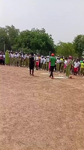 The video shows a group of people, likely students, gathered on a dusty, open field in Dutse, Nigeria. The sky is bright and overcast, suggesting daytime with potentially warm weather. A group of individuals in khaki uniforms and white shirts are lined up, possibly for an activity or event. In the foreground, a few individuals are standing and seemingly observing the group. One person in the foreground wears a red bandana and a red and white shirt, their back to the camera. Another person, wearing a black shirt and shorts, stands with their back to the group, facing the field. A person in a bright green outfit stands out among the uniformed individuals, and another person in a pink dress is also visible.

The scene appears to be an outdoor gathering or a sporting event. The sandy ground and the presence of a fence in the background suggest an organized space like a schoolyard or sports complex. The overall atmosphere seems to be one of anticipation or instruction, with some individuals appearing to be giving directions or observing participants. There is no visible text in the video.