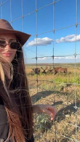 A young woman with long dark hair and blonde highlights, wearing a brown cowboy hat and sunglasses, stands in front of a wire fence. She is dressed in a black top. She smiles and extends her left hand towards the fence. Behind her, in a grassy field, several deer are visible. The sky is blue with scattered clouds. The scene appears to be outdoors in a natural environment, possibly a wildlife reserve or ranch.