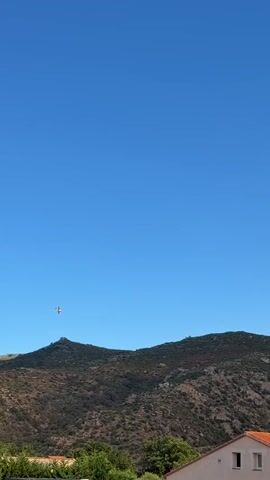 The video captures a wildfire occurring on a hillside under a clear blue sky. The fire is evident through a large plume of smoke rising from behind a ridge. An airplane is visible in the sky to the left of the smoke plume, appearing to be in the vicinity of the fire. The lower portion of the video shows a red-tiled roof of a building and green foliage, indicating a populated or inhabited area near the fire. The smoke is dense and appears to be expanding rapidly upwards and outwards.