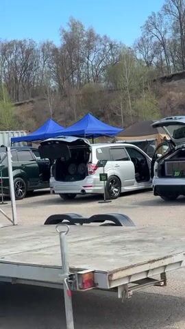 The video shows an outdoor car audio competition. Several cars are parked with their trunks open, revealing large audio systems inside. The cars include a silver Subaru Forester, a dark green SUV, and a white hatchback with a purple speaker box. Several blue canopies are set up in the background. A man in shorts and a t-shirt walks between the cars. A flatbed trailer with a license plate reading "7666 BO 25 RUS" is visible in the foreground. The location appears to be a gravel parking lot near a wooded area on a sunny day.