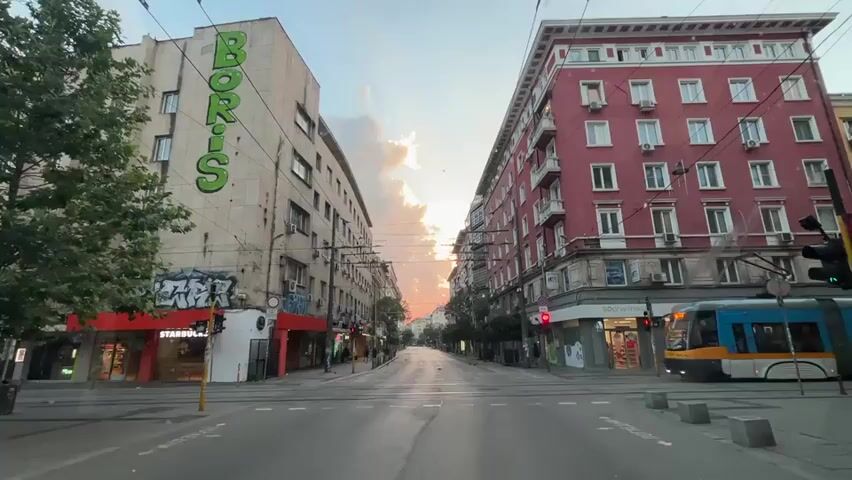 The video shows a street scene in a city at what appears to be dusk. On the left, a light-colored building has large green lettering spelling "BORIS" on its facade, along with some graffiti at its base. Below this is a store with a red awning, and the word "STARBUCKS" is visible. On the right, a red-colored apartment building with many windows rises several stories. A blue and yellow tram is moving from right to left across the frame. In front of the tram, a person on a bicycle is also moving from right to left. The street is paved and appears to be an intersection, with traffic lights and overhead wires for the tram visible. Trees are present on the left side of the street. The sky is partly cloudy with the sun setting, casting a warm glow. The overall impression is that of an urban environment during a transitionary time of day.