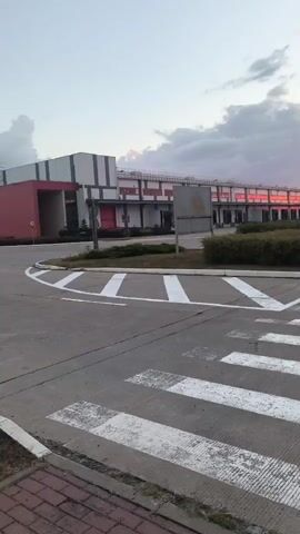 This video shows a wide shot of an industrial area with a large, modern building in the background. The building has a mix of grey and red sections, with many windows reflecting the colorful sky. There is a pedestrian crossing in the foreground, and the area is landscaped with bushes and trees. The sky is filled with dramatic clouds tinged with pink and orange hues, suggesting sunrise or sunset. The overall impression is one of a quiet, functional, and aesthetically pleasing industrial environment during a beautiful natural light display.