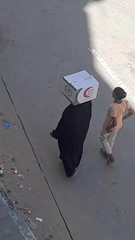 A person wearing a black abaya carries a white box on their head. A young boy in an orange shirt and beige pants walks beside them. The box has a red crescent symbol and Arabic writing on it. They are walking on a paved surface with some debris visible. Another person's legs are visible in the background.
