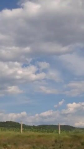 The video shows a view from a moving vehicle. The foreground is a blurry grassy field with some green bushes and trees. Two wooden fence posts are visible. In the middle ground, there is a line of trees and hills. The sky is filled with white and gray clouds against a blue background. The camera pans slightly to the right.