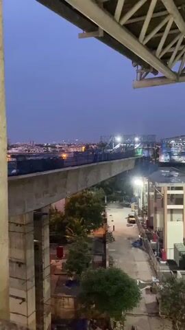 This video captures a construction scene at dusk, likely in the St. Thomas Mount-Pallavaram Cantonment Board area of India. The primary subject is an elevated structure, possibly a bridge or flyover, under construction. A long, concrete beam forms the foreground, extending diagonally across the frame.

Above the beam, workers in orange vests are visible, engaged in ongoing construction activities. They are silhouetted against the dimming sky and illuminated by temporary work lights. The structure itself is a complex network of scaffolding, metal beams, and blue tarpaulin sheeting, indicating a significant civil engineering project.

The setting appears to be an urban or semi-urban environment. In the background, a city skyline with buildings and lights can be seen through the twilight haze. Trees and foliage are present below the elevated structure, suggesting a mix of developed and natural areas. The ground level appears to be a construction site or access road, with a yellow vehicle and some construction materials visible.

The time of day is clearly dusk or early evening, indicated by the deep blue sky and the artificial lighting dominating the scene. The weather appears to be clear, with no visible rain or strong winds. The overall mood is one of active work continuing into the evening hours, characteristic of construction projects that aim to meet deadlines. No visible text is present in the video.