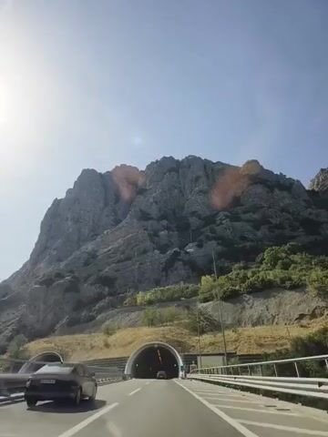 The video shows a first-person perspective of driving on a highway. In front of the car, a tunnel is visible with lights inside. A large, rocky mountain looms behind the tunnel. The sun is shining brightly, causing some glare in the video. There is a black car in the lane to the left of the driver. Guardrails are visible on the side of the road.