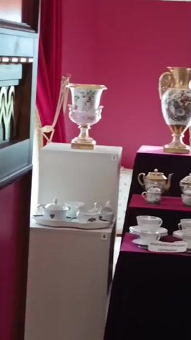 The video shows an exhibition of porcelain items in a room with pink walls and red curtains. On a white pedestal on the left is a porcelain vase with a floral design, and below it is a porcelain tea set on a tray, labeled with the number 4. To the right of the first pedestal is another display with items arranged on tiered shelves. The top shelf holds two small teapots and two covered sugar bowls, labeled with the number 5. The middle shelf has four teacups and saucers, and the bottom shelf has four more teacups and saucers, with a label that reads "ИМПЕРАТОРСКИЕ СЕРВИЗЫ." The second display from the left has a large gold vase with a floral design on top, and a smaller porcelain vase with a floral design below it, labeled with the number 1. To the right of this display is another white pedestal with a porcelain vase with a floral design and a gold base. Further to the right is a tall, narrow porcelain vase with a floral design on a gold base, placed near a window with white curtains and red draperies.