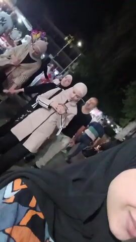 The video shows a group of people standing on what appears to be a street or public area at night. There are several women wearing hijabs, one wearing a light-colored coat with black stripes. A man is standing nearby. Other people can be seen in the background, along with a car and some street infrastructure. The camera is held by someone who seems to be part of the group, capturing a low-angle view of the scene. The overall lighting is dim.