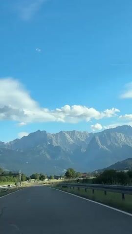 This is a vertical video clip of a scenic drive through a mountainous region, likely in Bavaria, Germany, given the location context of Farchant. The camera is positioned inside a vehicle, looking forward as it travels along an asphalt road.

The primary subjects are the majestic mountains that dominate the background. They are rugged and appear to be a range of peaks, with some visible snow or ice caps, suggesting a higher altitude. The slopes leading up to the mountains are covered in dense green forests.

In the midground, there are signs of rural life. Green fields stretch out on either side of the road, and scattered amidst them are a few trees. A small village or hamlet is visible in the distance, with a church steeple and red-roofed buildings. A guardrail runs along the right side of the road, and some road signs are present.

The sky is a vibrant blue with scattered fluffy white clouds, indicating fair weather. The lighting suggests it is daytime, possibly late morning or early afternoon, as the sun is high enough to cast clear shadows and illuminate the landscape brightly. The overall mood is peaceful and evokes a sense of adventure and natural beauty. There is no visible text in the video.