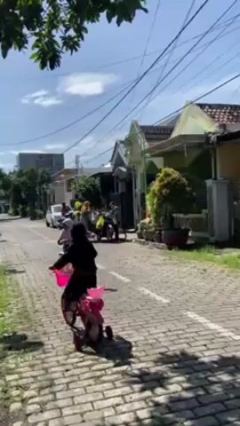In a neighborhood on a bright day, two girls are riding bicycles on a paved street. The street is lined with houses and trees. One girl is wearing a white hijab and a green dress. She is riding a pink bicycle. The other girl is wearing a black outfit and is also riding a pink bicycle. In the background, adults in hijabs stand near a vehicle. Overhead, electrical lines stretch across the sky.