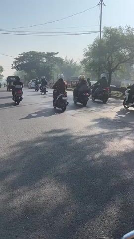 A group of motorcyclists are riding down a paved road in a convoy. The motorcyclists are wearing helmets and some are carrying passengers or cargo. The road is lined with trees and there are some buildings visible in the background. The sun is shining and the sky is clear. The video is shot from the perspective of another motorcyclist.