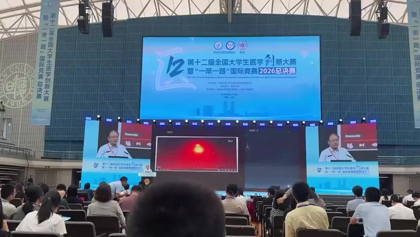 The video captures a scene from an event held indoors, likely a conference or competition, with a large screen displaying visuals and text. The screen prominently features Chinese characters and English text that reads "The 12th China Undergraduate Medical Innovation Tournament & The Belt and Road International Competition 2026 Final". There are two speakers, one on the left and one on the right side of the stage, both appearing to be male and dressed in formal attire, possibly giving presentations or speeches. They are positioned behind podiums. Between the speakers, a large screen displays a video feed, featuring what appears to be a red background with small, bright red dots, possibly representing data or a simulation. Below this main screen, a smaller screen also shows a similar red visual with the text "2025" above it, and another screen on the right shows fireworks-like visuals with the text "2026". The audience is seated in rows facing the stage, with many people in the foreground, some looking towards the stage and others looking down. The environment appears to be a large hall, with stage lighting and a muted background wall. The overall atmosphere suggests a formal academic or professional event.