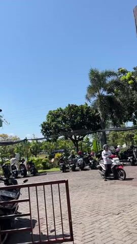 The media file depicts an outdoor scene under a clear blue sky, suggesting a bright, sunny day. The setting appears to be a parking area, possibly outside a business or attraction, paved with interlocking bricks. Lush green trees and foliage surround the area, with palm trees visible in the background, hinting at a tropical or subtropical climate.

Several motorcycles are parked and in use. In the foreground, a red metal barrier is visible. A woman wearing a white outfit and a helmet is riding a pink and white motorcycle, moving from right to left across the frame. Other motorcycles are parked in rows further back, with a few individuals near them, though their activities are less clear. One person is standing near a parked motorcycle on the left, wearing light-colored clothing.

The time of day is likely midday due to the bright sunlight and long shadows. The overall atmosphere seems relaxed and ordinary, with no strong emotional cues apparent. The location context provided is Pagerwojo, Indonesia, which aligns with the visual cues of tropical vegetation.

There are some visible text elements on license plates of the motorcycles. One license plate on the left side reads "AB 2008 AE", and another on a parked motorcycle in the background appears to have characters that are not clearly legible. The scene captures a moment of everyday activity in what seems to be a public outdoor space in Indonesia.