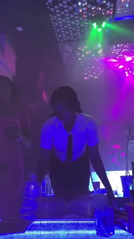 A group of people are in a dimly lit club. The lighting is a mix of purple and green, with disco ball-like reflections on the ceiling. A woman in a white shirt and dark shorts is leaning over a table. There are drinks on the table, including a bottle of water and a red cup. Another woman in a pink shirt is visible on the left side of the frame. The overall atmosphere is that of a party or celebration.