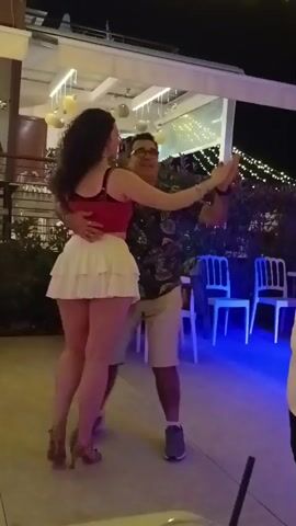 A man and a woman are dancing outdoors at night. The woman is wearing a red top and a white skirt. The man is wearing a patterned shirt and tan shorts. They are holding each other and appear to be dancing to music. There are tables and chairs in the background, as well as string lights.