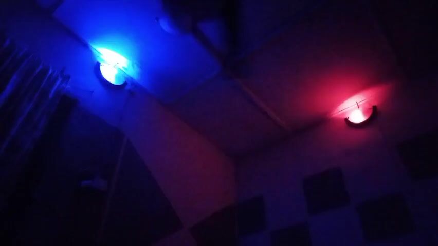 The video shows a room corner with two lights: one blue and one red, illuminating the space. The blue light is on the left side of the frame, and the red light is on the right. The walls are decorated with what appears to be soundproofing panels. A curtain hangs partially visible on the left side. The ceiling appears to be comprised of square tiles. The overall lighting is dim.