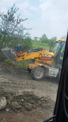 This video shows a yellow JCB backhoe loader operating on a muddy, uneven terrain. The machine is seen from inside another vehicle, likely a cab, as evidenced by the framing and the presence of a black belt and what appears to be part of a dashboard in the foreground. The backhoe is actively digging or moving material with its front bucket, kicking up dust and mud.

In the background, there is a line of trees and some distant structures, possibly industrial or agricultural. The sky is overcast, suggesting a cloudy day. The overall scene depicts construction or ground-clearing work in a rural or semi-industrial setting in Novyi Rozdil, Ukraine. No people are clearly visible within the backhoe's cab, though a figure might be present. The activity is focused on the operation of heavy machinery. Visible text on the backhoe includes the "JCB" logo and model numbers like "3CX".