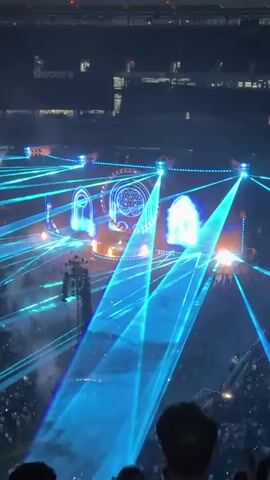 An aerial view of a concert stage with a large screen displaying abstract circular imagery and faux stained-glass windows. Blue laser lights crisscross the arena, illuminating a massive crowd of concertgoers. The stage itself features spotlights and what appear to be pyrotechnic effects. A large speaker system is visible on a raised platform to the left of the stage. The lighting transitions from blue to purple, and the crowd is a dark mass of silhouettes.