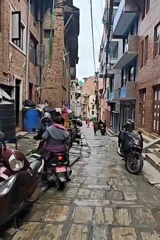 The video shows a narrow, cobblestone street in a city. Buildings line both sides of the street, with many of them appearing to be older brick structures. There are several motorbikes parked along the sides of the street. In the foreground, a person is sitting on a motorbike, facing away from the camera. Further down the street, a few people are walking. Wires are strung overhead, indicating a developed urban area. The street appears to be wet, suggesting it may have recently rained or is about to rain. The overall atmosphere is one of a bustling, lived-in urban environment.