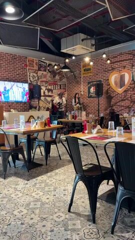 The video captures a lively scene inside a restaurant. In the background, a duo performs music; a man plays an electric guitar and sings, while a woman stands beside him, also singing and playing a drum. They are positioned on a small stage area in front of a brick wall adorned with vintage-style posters featuring cars and advertisements, including one for "MOTOR OIL." The restaurant's interior features industrial-style lighting fixtures and a distinctive patterned floor. In the foreground, several tables are set with red placemats, cutlery, glasses, and salt and pepper shakers, suggesting a dining setting. A television screen is visible on the left side of the frame, displaying colorful content. The overall atmosphere appears to be energetic and casual, with the live music adding to the ambiance.