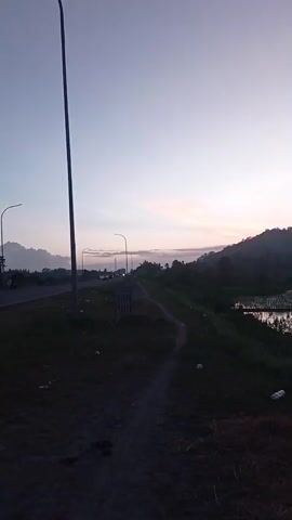 The video opens with a tall, dark street lamp on the left side of the frame. A paved road with street lights stretches into the distance on the left. A dirt path runs along the top of a grassy embankment. On the right side of the embankment, there is a body of water, possibly a rice paddy, reflecting the sky. In the background, there are hills covered in vegetation. The sky is a pale gradient of light blue and pink, suggesting either sunrise or sunset. The overall scene is peaceful and rural. The video pans slowly to the right, revealing more of the landscape and the reflection in the water.