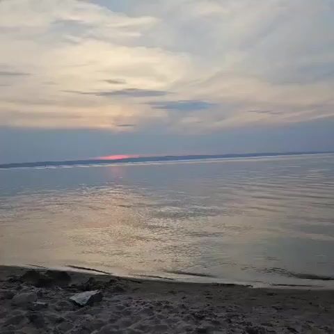 The video shows a natural landscape with a body of water and a shoreline. The sky is filled with clouds, some of which are tinged with pink and orange hues, suggesting it is either sunrise or sunset. The water is calm with gentle ripples. The shoreline in the foreground is sandy and appears to have some debris on it. The overall scene is serene and peaceful. There are no people or animals visible in the video.