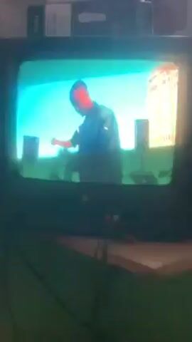 An old, black, boxy television is playing a music video of a black man rapping. The video starts with a wide shot of the man standing on a sunny city rooftop. It then cuts to a green filter, followed by a shot of the man directly facing the camera, clapping. Above the television is a white cable box and what appears to be a black video game console. A green wall sits behind the television.