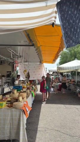 A busy outdoor market is depicted, with a focus on a cheese stall.  Two customers, a woman in a red dress and a man in a green shirt and shorts, are interacting with a vendor at the cheese stall. The woman is pointing at something on the stall. The stall itself is laden with a variety of cheeses, some whole wheels and some cut portions.  The vendor, a woman in a white top, is behind the stall.  The stall is covered by a striped awning.  In the background, other market stalls and shoppers can be seen.  The ground is paved.