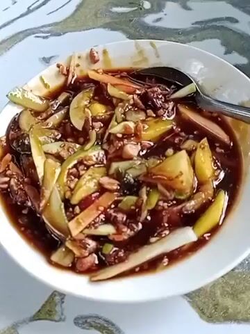 A close-up shot shows a white bowl filled with a mixed fruit salad. The salad appears to be made with thinly sliced fruits such as cucumbers, mangoes, and possibly apples, all coated in a dark, thick sauce. Scattered throughout the salad are chopped peanuts and some darker, possibly dried, ingredients. A silver spoon rests in the bowl, partially submerged in the sauce. The bowl is placed on a table with a patterned surface, likely a tablecloth. The lighting suggests it is daytime, possibly indoors.