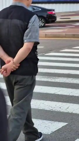 Two individuals are walking across a pedestrian crossing. The person in the foreground is wearing a dark vest over a light gray short-sleeved shirt and dark gray pants. Their hands are clasped behind their back. The person behind them is wearing a blue polo shirt and light gray pants. In the background, a woman with dark hair wearing a plaid shirt, a brown baseball cap, and a backpack is walking while looking at her phone. She is also crossing the street. The environment appears to be a street with buildings and storefronts, including signs for SK Telecom and KT.