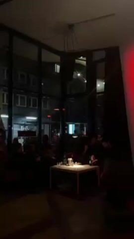 The video shows a dimly lit indoor space, possibly a bar or lounge, at night. Several people are seated around a low white table, which holds various bottles, possibly drinks, and a lit candle. The individuals appear to be engaged in conversation or simply relaxing. The room features large glass windows that provide a view of the street and buildings outside. A speaker is visible hanging from the ceiling. A red light is present on the right side of the frame, adding to the dim atmosphere.