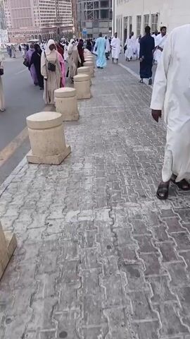 The video shows a sidewalk in what appears to be a city, with a crowd of people walking in both directions. Many individuals are wearing traditional Muslim attire, including hijabs and white robes. The sidewalk is paved with interlocking concrete blocks and lined with short, cylindrical concrete bollards. On one side of the sidewalk is a low, white building, while on the other side is a road with more pedestrians. Tall buildings are visible in the background, some of which are under construction. The camera appears to be held by someone walking along the sidewalk, capturing the scene as they move.